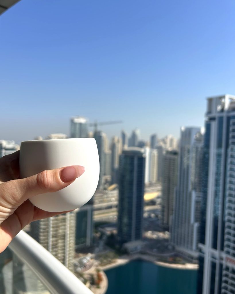 A white ceramic coffee cup held against the Dubai Marina skyline from a high balcony, towers rising into a clear blue sky.
